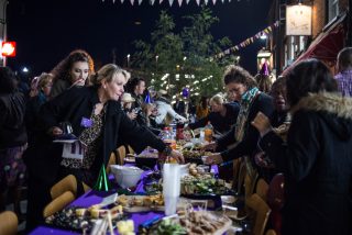Residents of Luton enjoying the street feast launching the public realm arts project. Image shows people gathered around a purple table laden with nibbles.