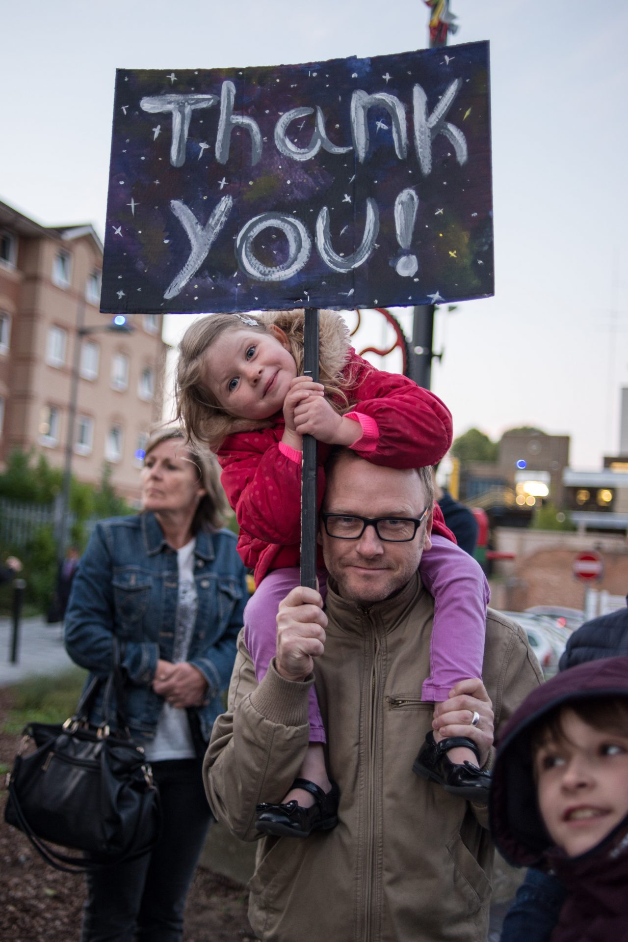 A father with his daughter on his shoulders, holding a placard that reads 'Thank You!' as part of Scott King's 'Keep the Home Fires Burning.'