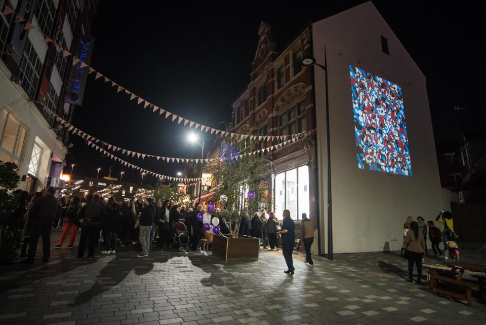 A brightly coloured projection of geometric shapes onto the Culture Wall at the launch of the public realm arts project with guests enjoying a feast next to it.